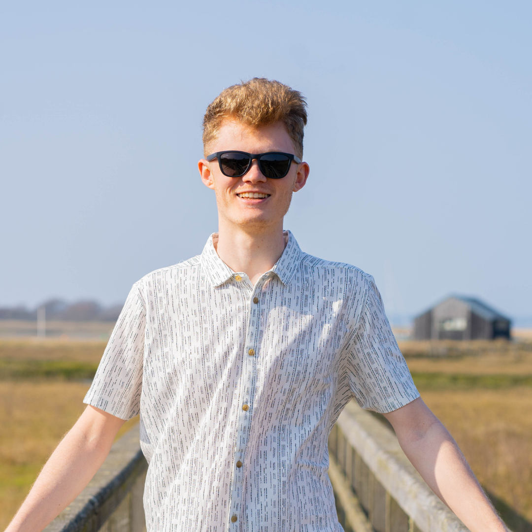 A person standing in a white and black patterned shirt wearing sunglasses smiling on a wooden boardwalk